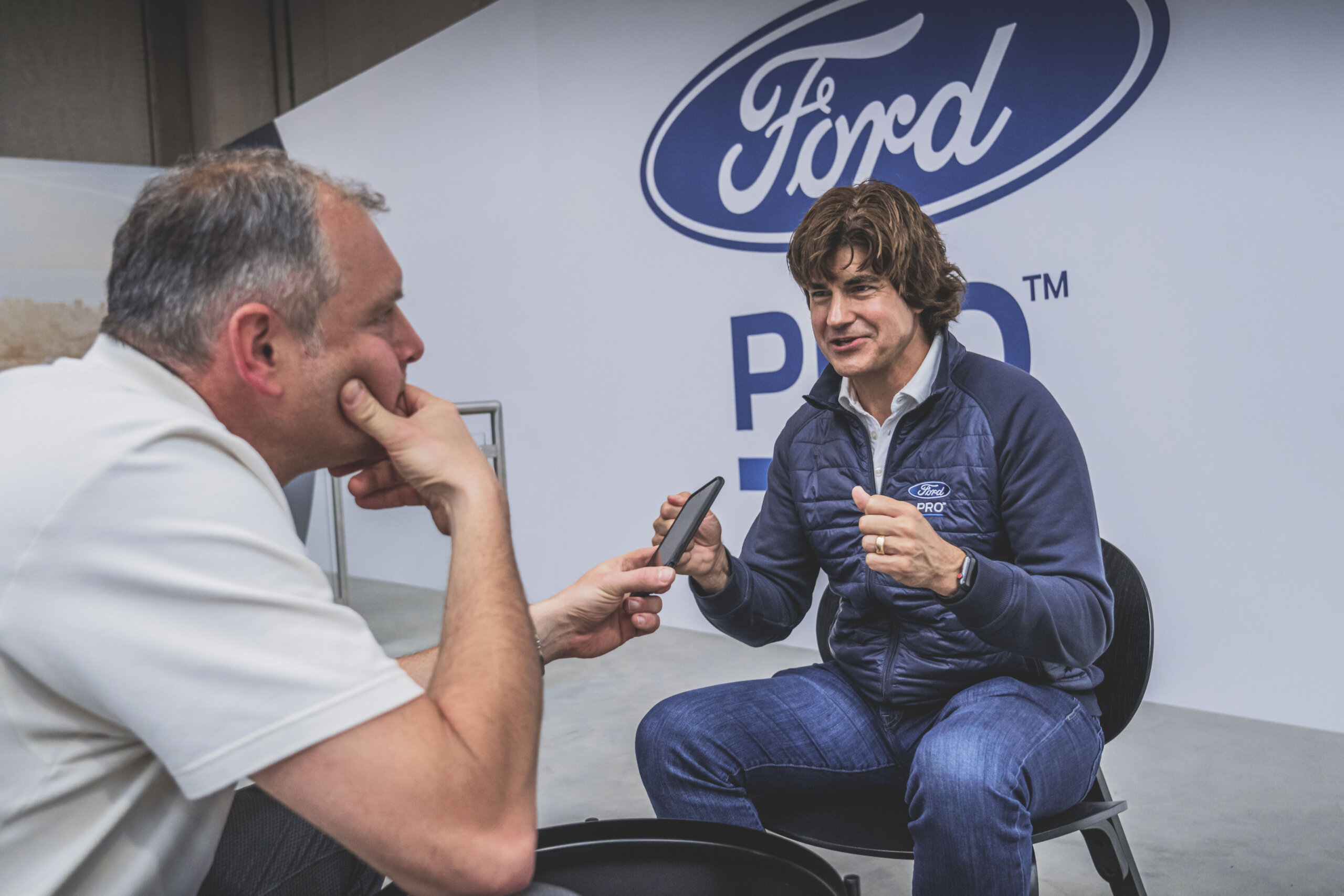 Dwóch mężczyzn rozmawia przed dużym logo firmy Ford; jeden z nich trzyma telefon.