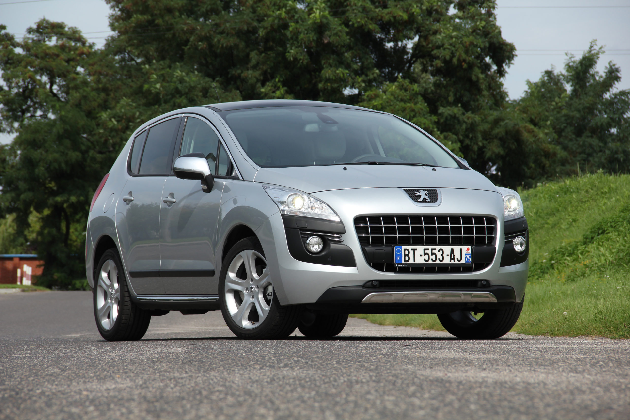 Srebrny SUV Peugeot 3008 stoi na jezdni, a za nim widać drzewa i francuską tablicę rejestracyjną.