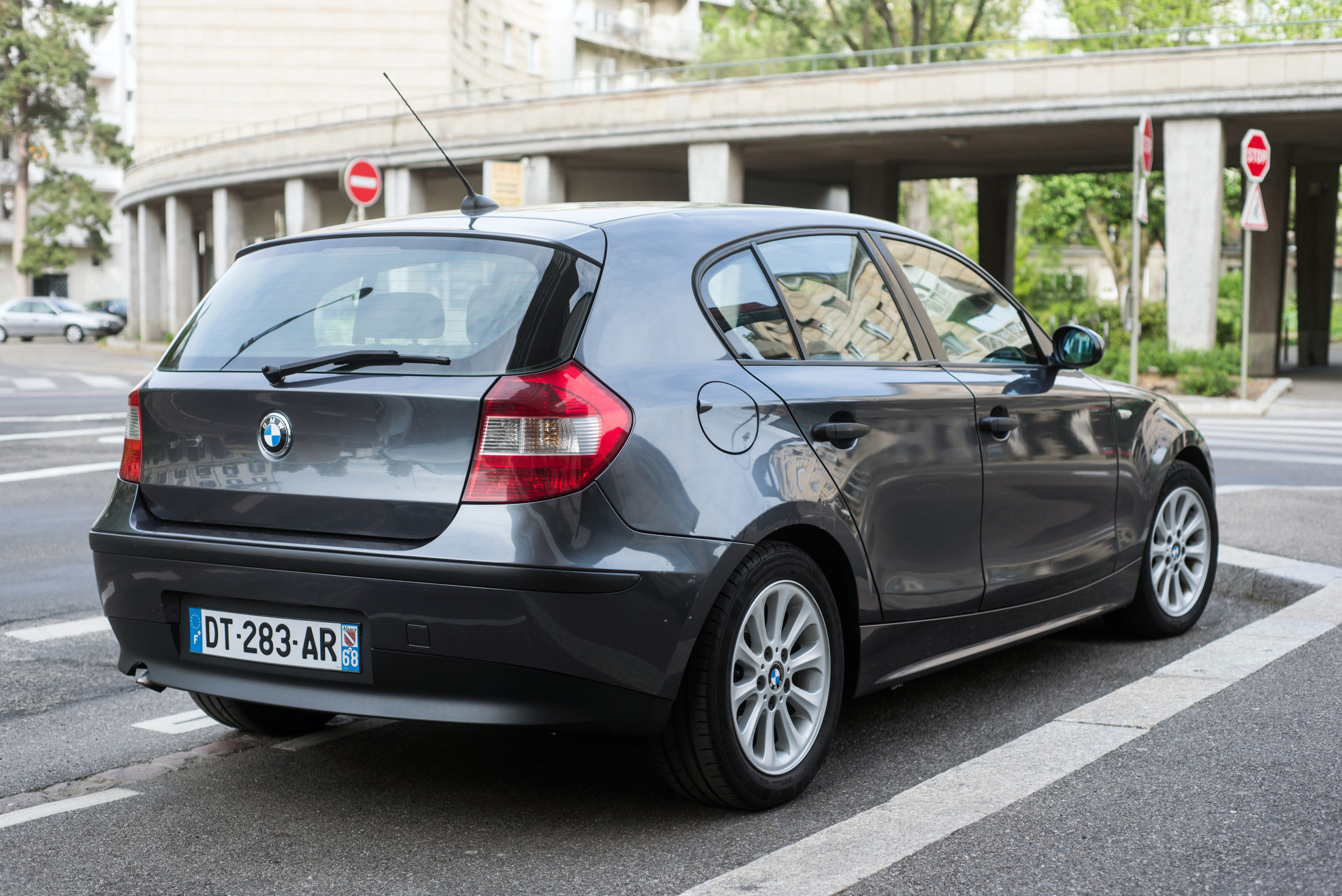 Szary hatchback BMW serii 1 zaparkowany przy rondzie, w tle budynki i zieleń.