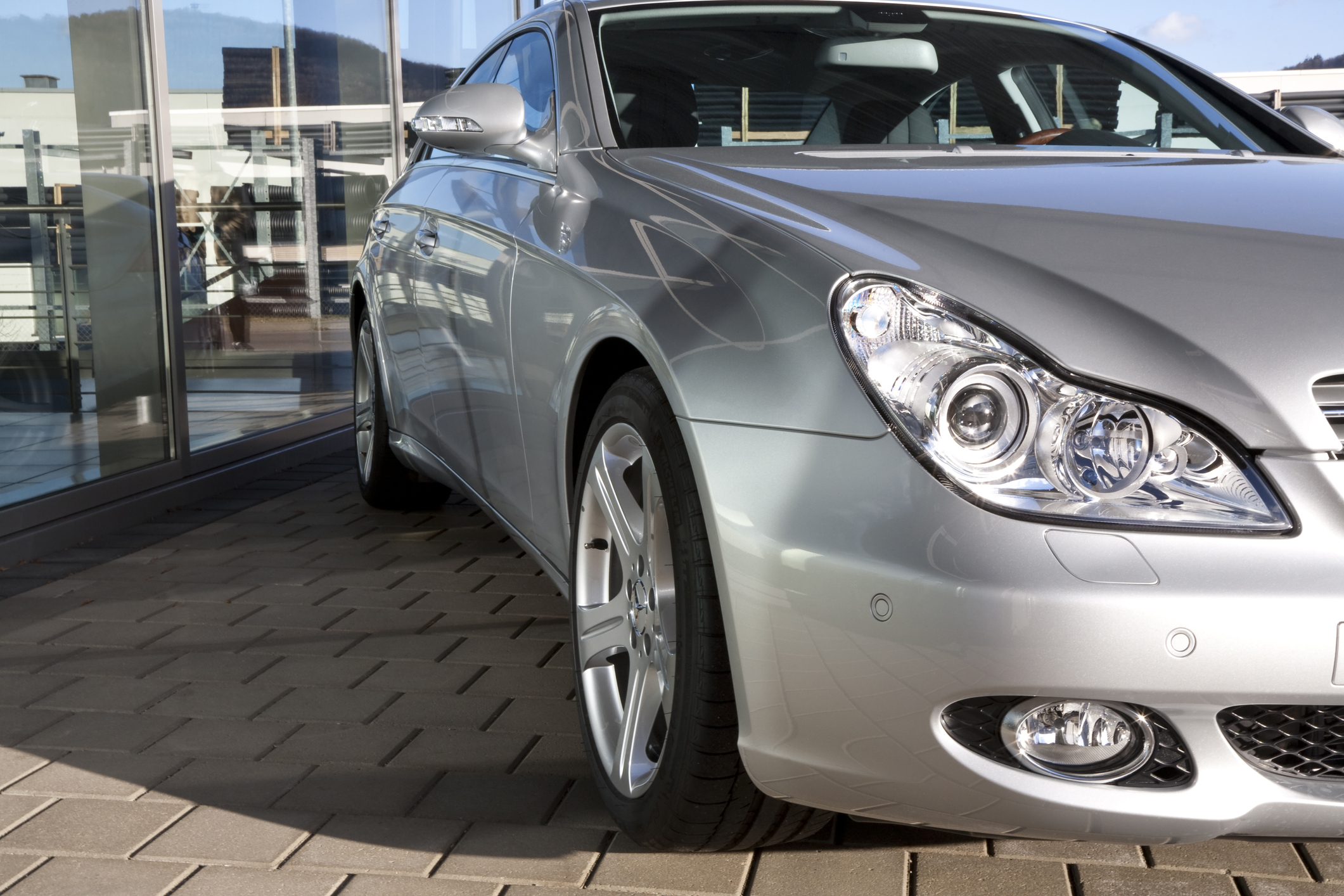 Srebrny Mercedes CLS na kostce przy szklanym budynku; widać bok, przód i koło.