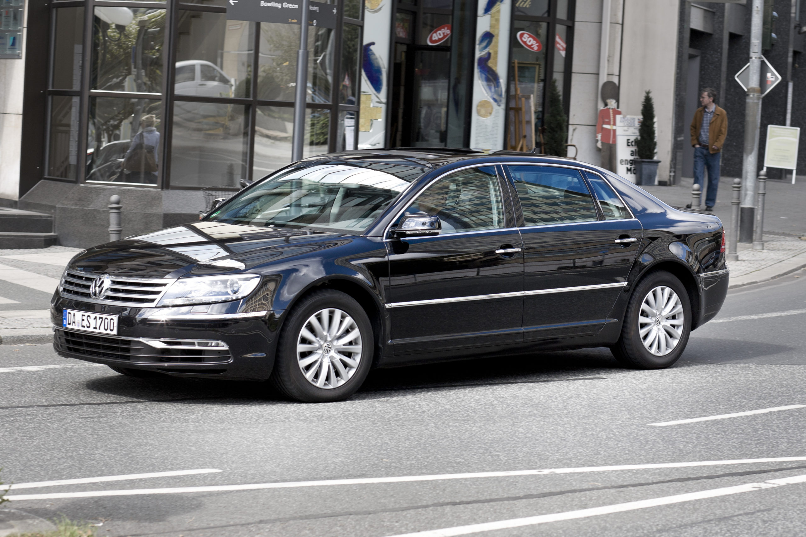 Czarny sedan marki Volkswagen, warty około 20 tys. zł, jedzie ulicą obok przeszklonego budynku.