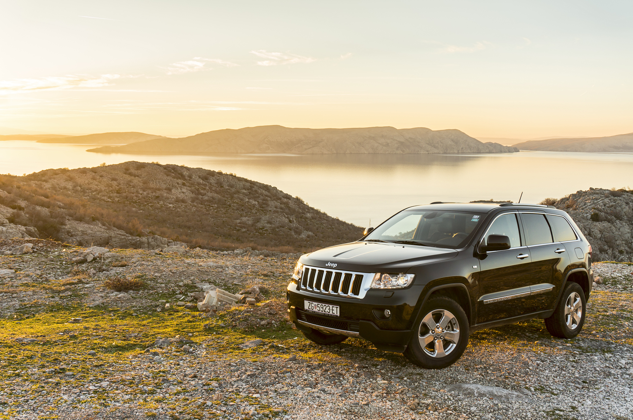 Czarny Jeep Grand Cherokee stoi na kamienistym terenie nad wodą o zachodzie słońca.