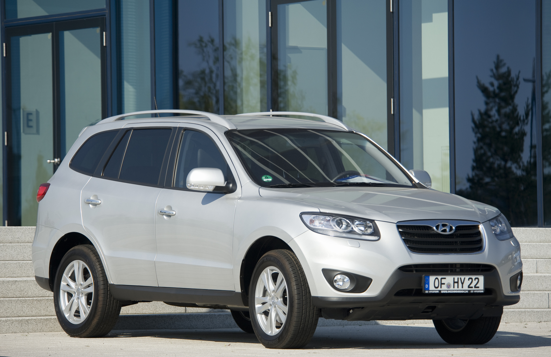 Srebrny Hyundai Santa Fe stoi na chodniku przed nowoczesnym budynkiem z szklanymi drzwiami.