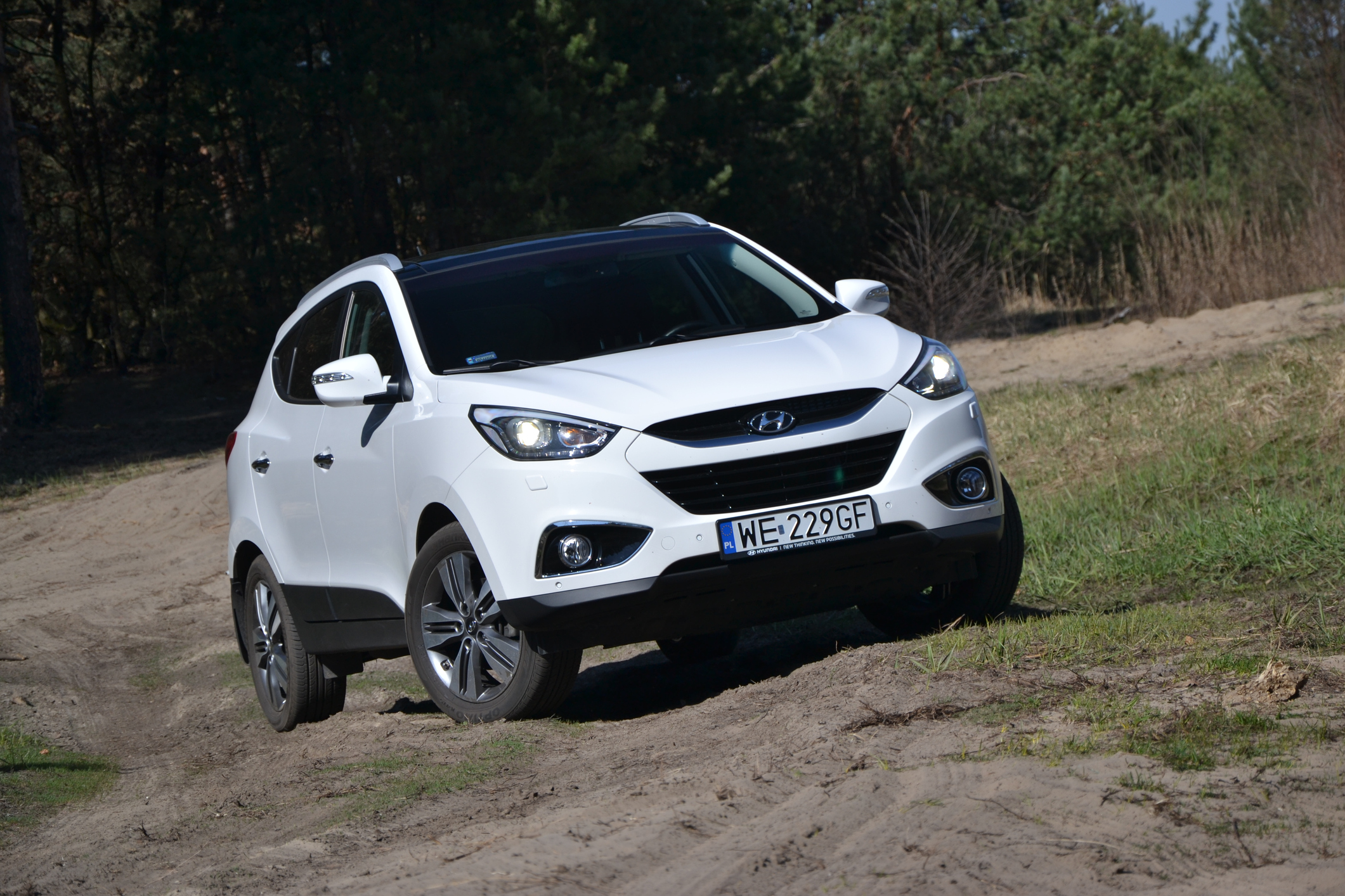 Biały Hyundai ix35 jedzie polną drogą wzdłuż trawy i drzew; tablica rejestracyjna WE 229GF.