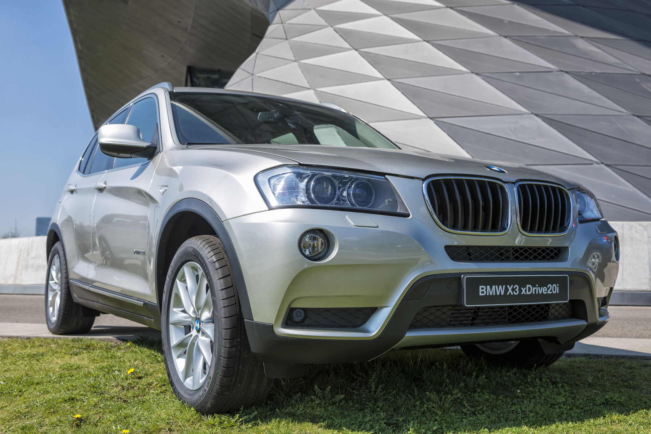 Srebrne BMW X3 xDrive20d stoi na trawie przed nowoczesnym budynkiem o geometrycznej architekturze.