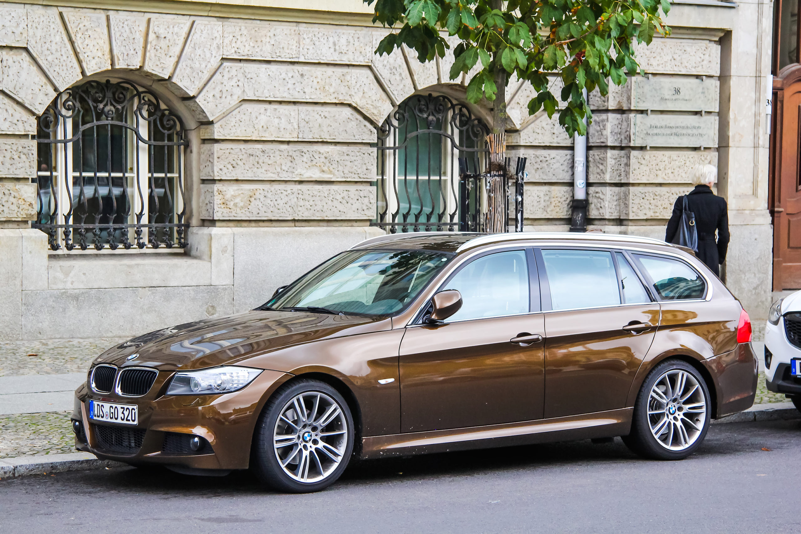 Brązowe BMW serii 3 E90 kombi stoi przy kamienicy, a osoba ubrana na czarno podchodzi do drzwi.