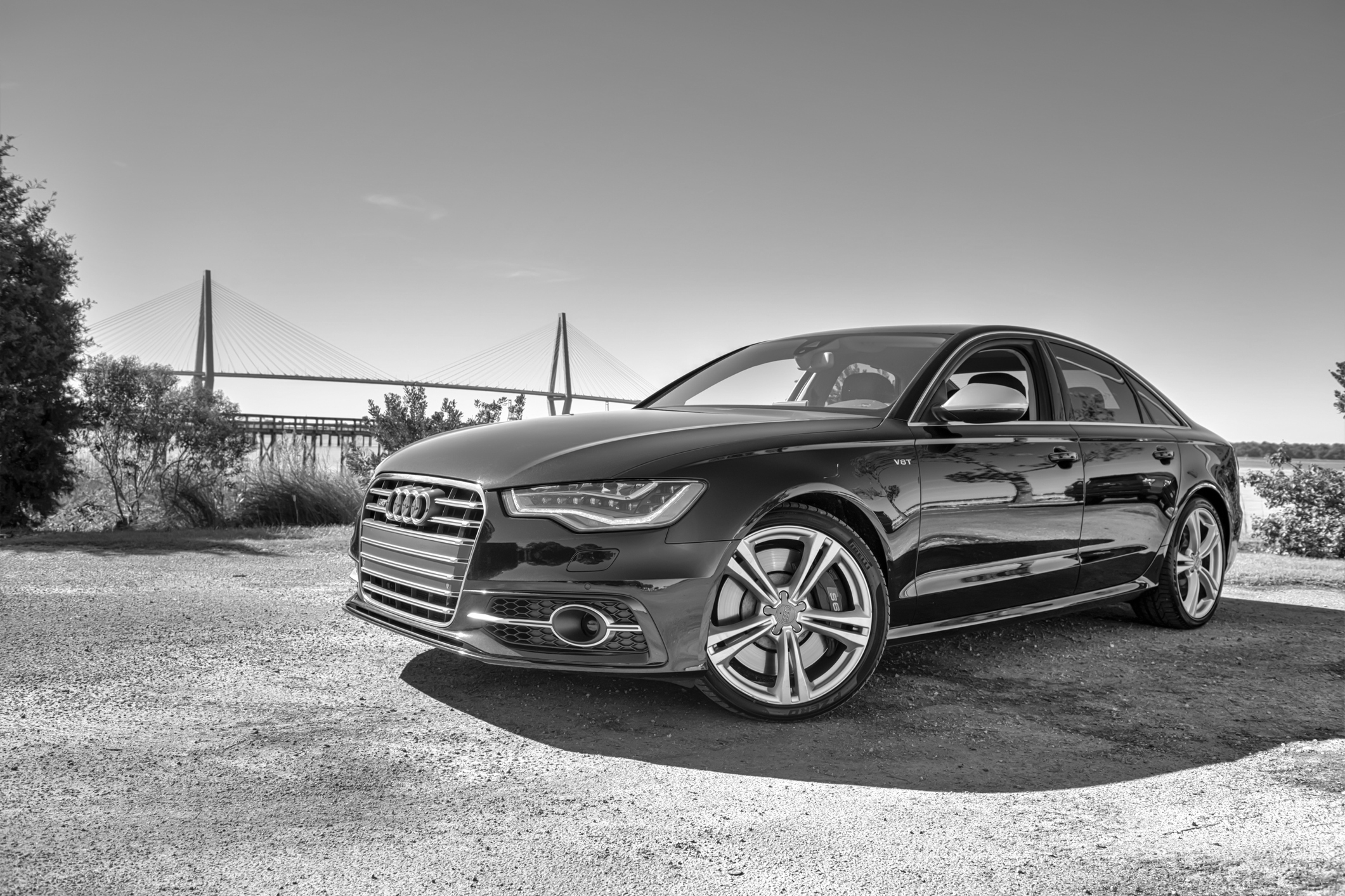 Czarno-białe Audi S6 stoi na polnej drodze, a w tle widać wiszący most i drzewa.