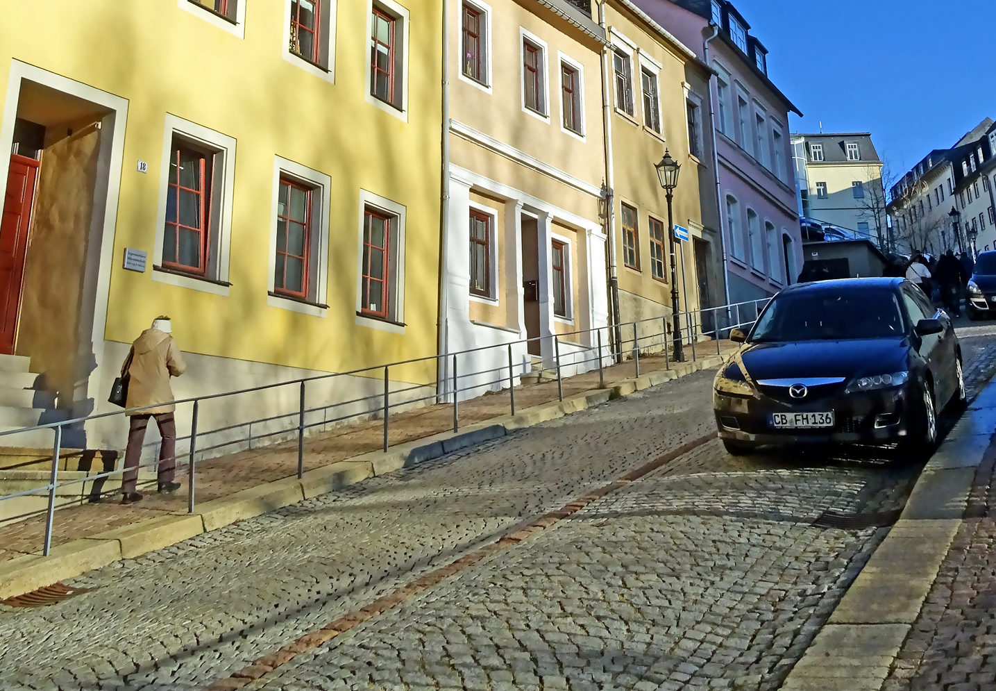 Brukowana ulica z Mazdą 6 i żółtymi budynkami; osoba idzie chodnikiem pod górę.