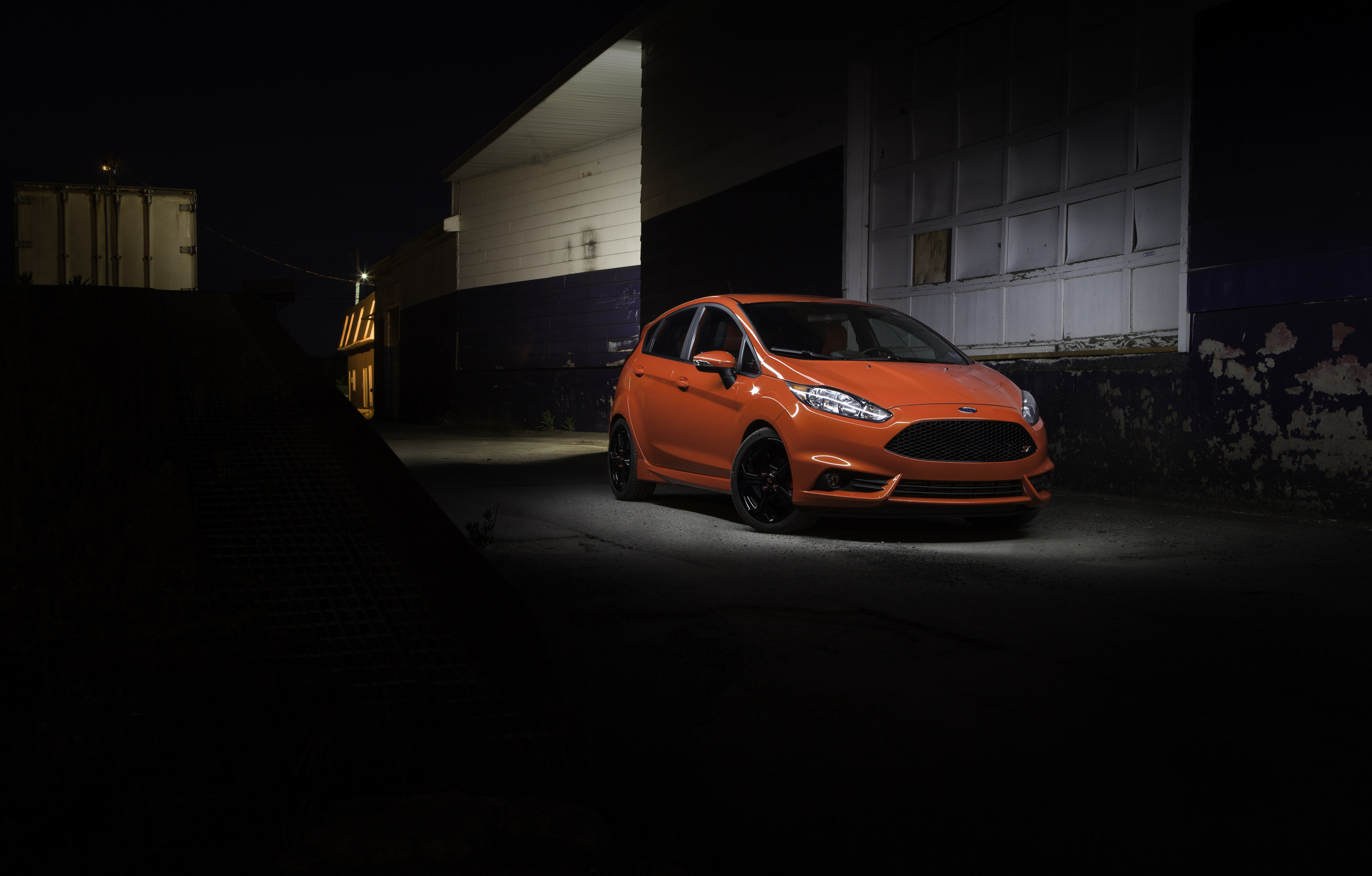 Pomarańczowy Ford Fiesta mk7 stoi nocą pod latarnią obok magazynu.