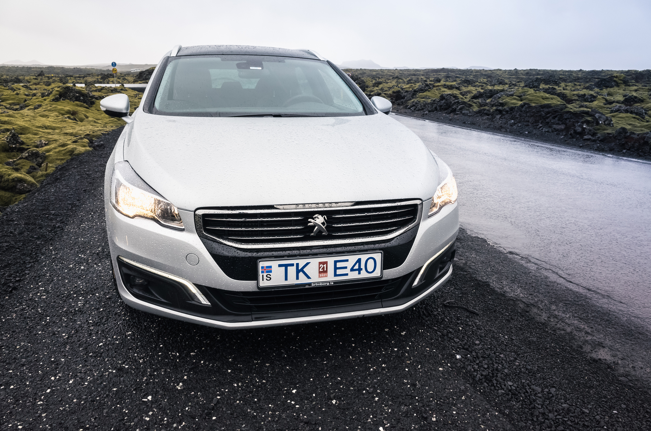 Srebrny Peugeot 508 z islandzkimi tablicami rejestracyjnymi TK E40 stoi na mokrej drodze obok skał.