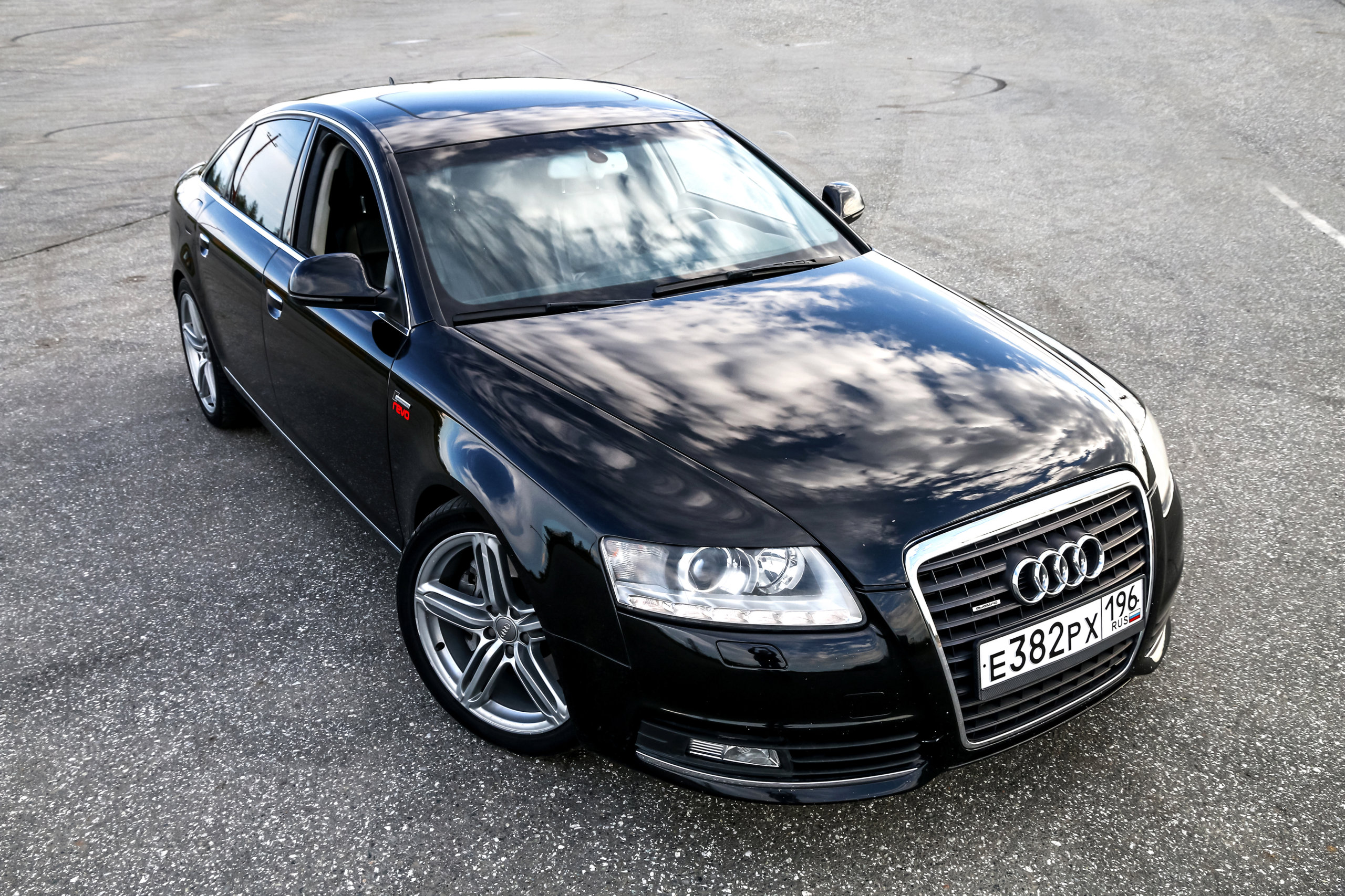 Czarny sedan marki Audi z rosyjską tablicą rejestracyjną stoi na pustym asfalcie, w którym odbija się niebo.
