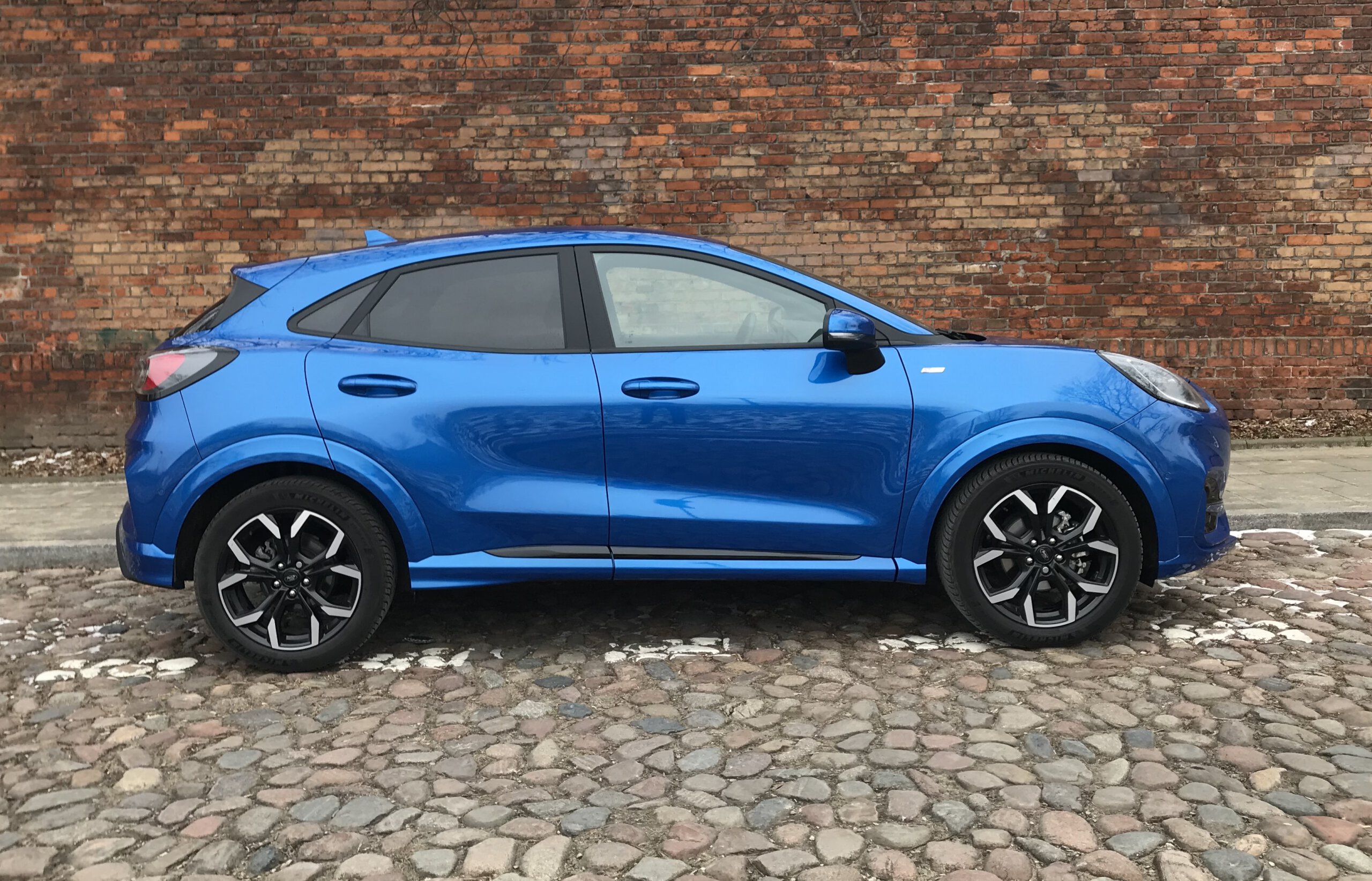 Niebieski Ford Puma stoi na brukowanej ulicy przed czerwoną ceglaną ścianą.