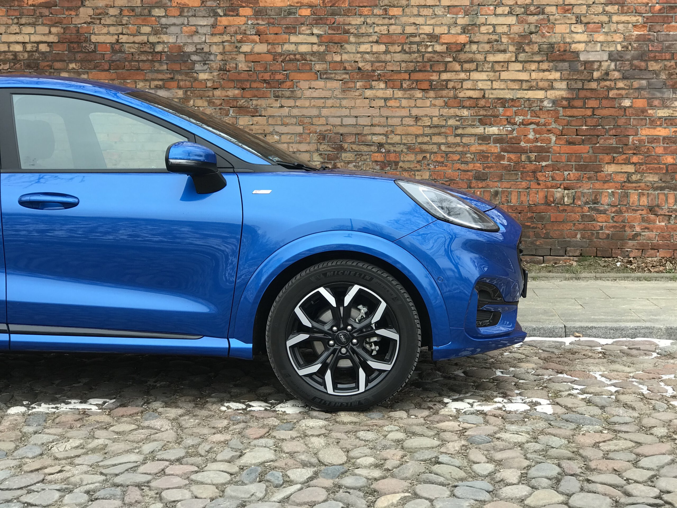 Niebieski Ford Puma zaparkowany przodem na brukowanej ulicy przy starej ceglanej ścianie.