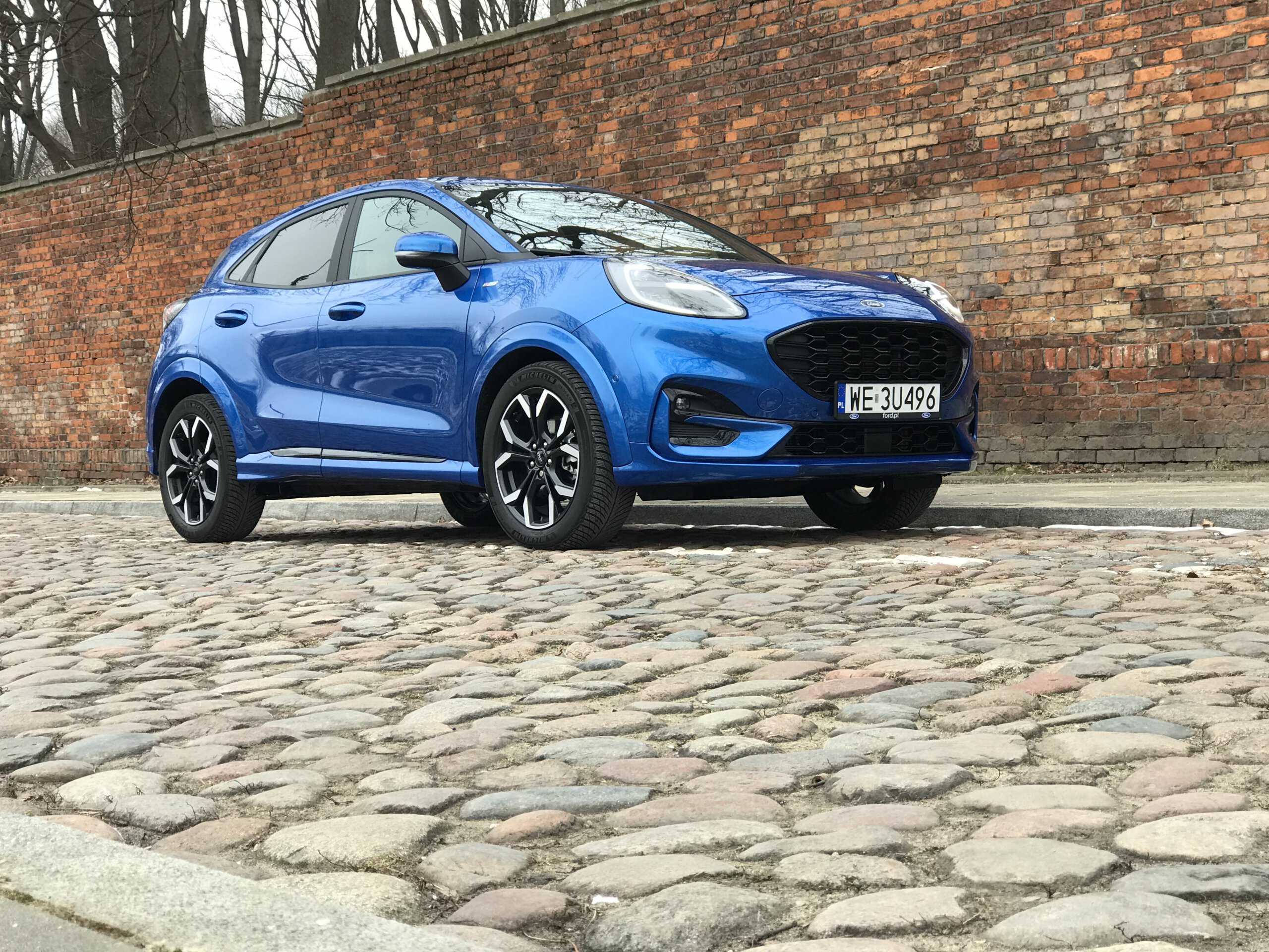 Niebieski Ford Puma stoi na brukowanej ulicy przed ceglanym murem i nagimi drzewami.