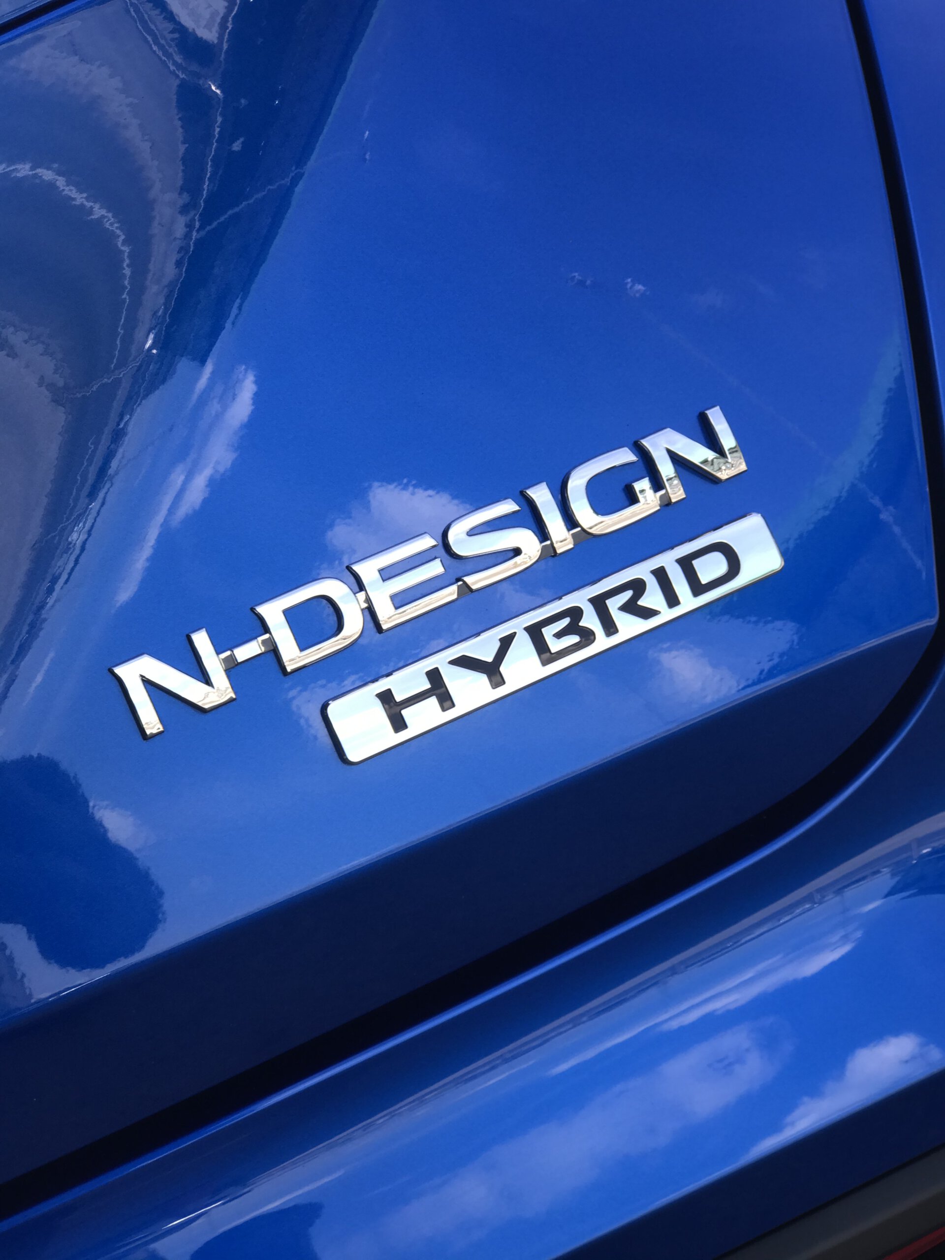 Zbliżenie na niebieskiego Nissana Juke Hybrid z chromowanymi emblematami N-DESIGN i HYBRID.