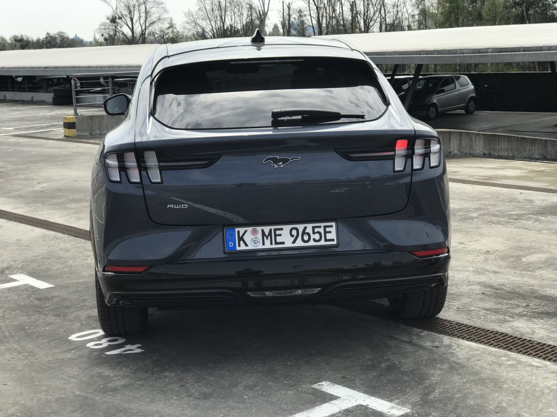 Szary Ford Mustang Mach-E z tablicą rejestracyjną K ME 965E stoi tyłem na parkingu pod drzewami.
