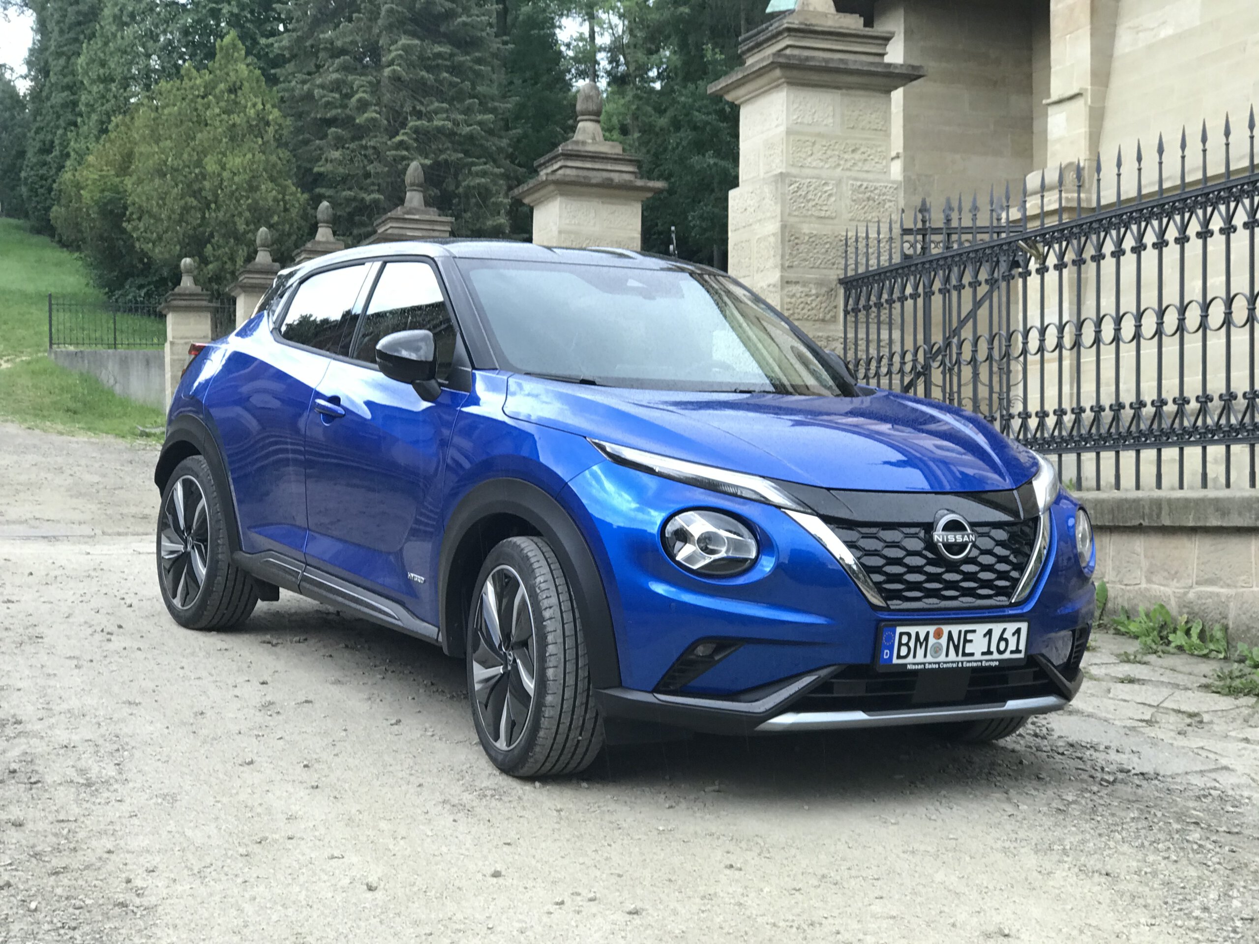 Niebieski Nissan Juke Hybrid stoi na polnej drodze przy czarnym ogrodzeniu i kamiennym budynku.