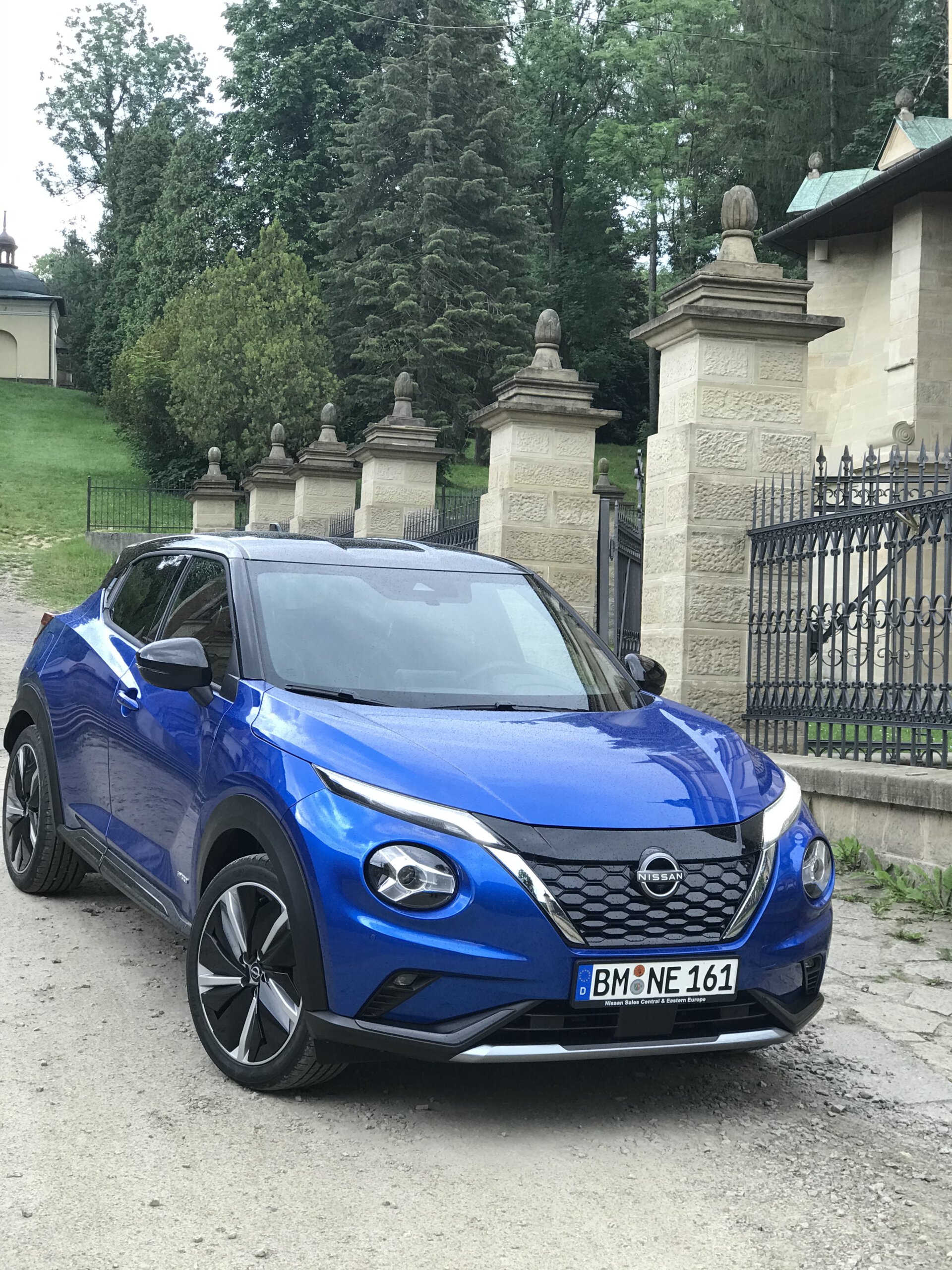 Niebieski Nissan Juke Hybrid stoi na polnej drodze obok kamiennego muru i zieleni.