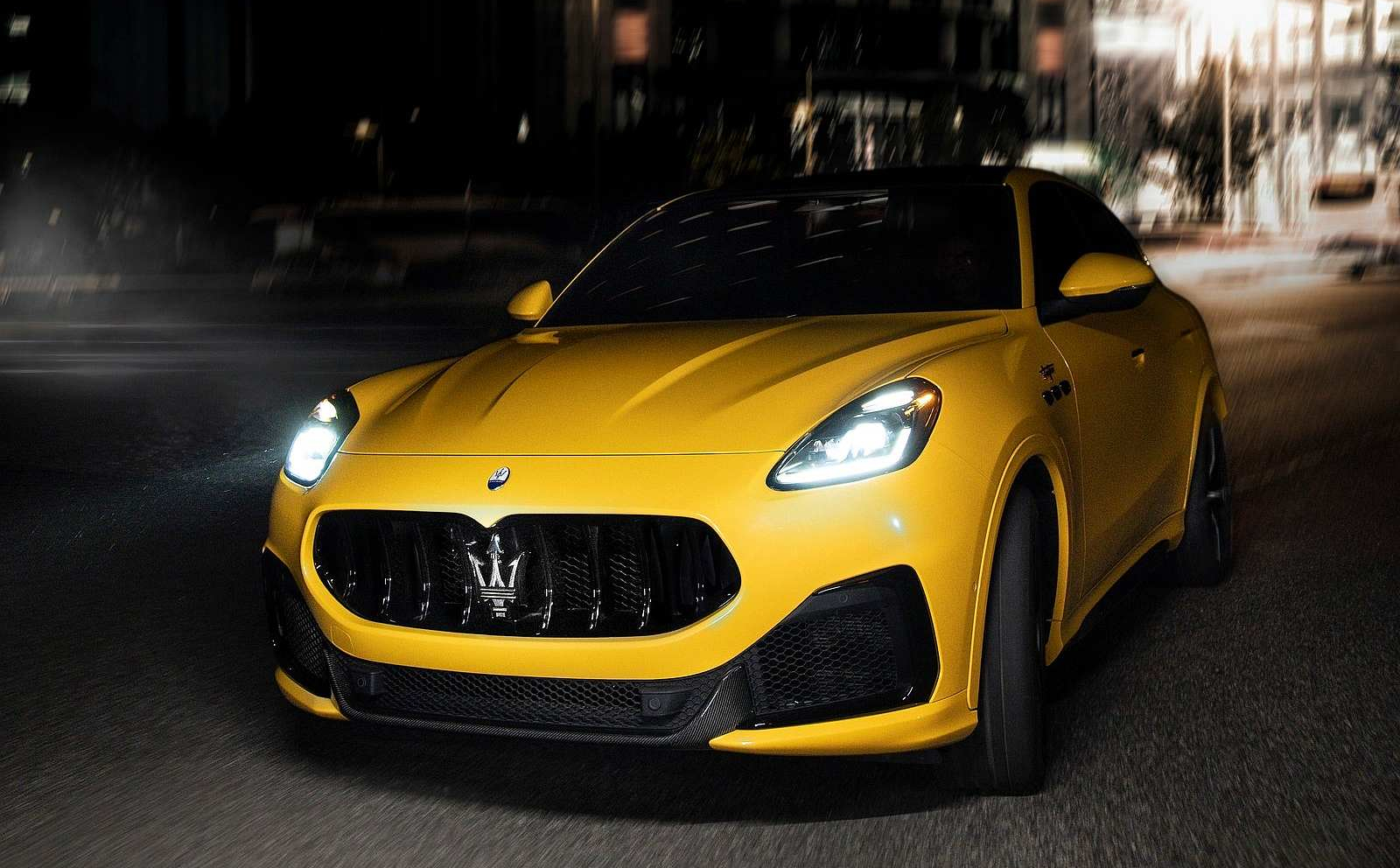 Żółte Maserati Grecale jedzie nocą ulicą miasta; światła oświetlają drogę, a budynki są rozmazane.