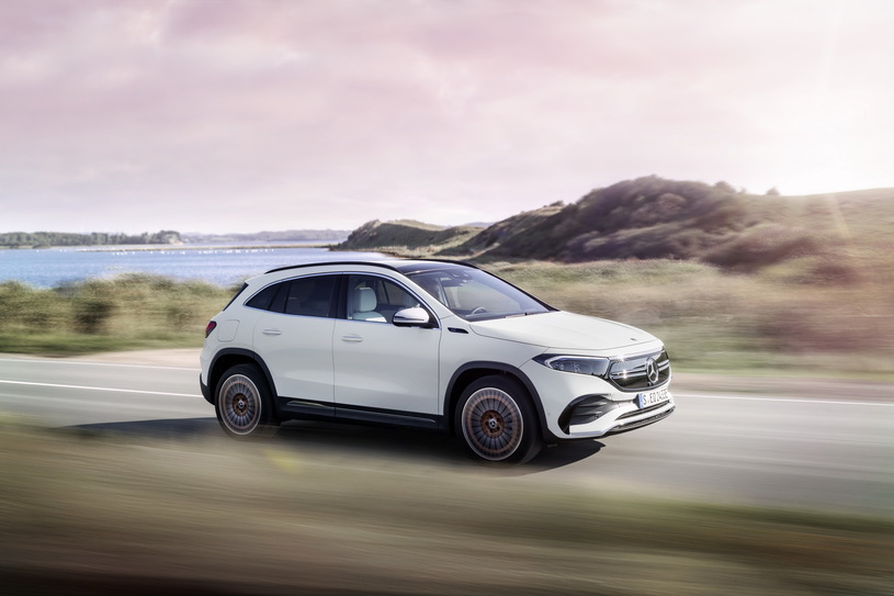 Biały Mercedes EQA jedzie drogą wzdłuż trawy i wody pod częściowo zachmurzonym niebem.