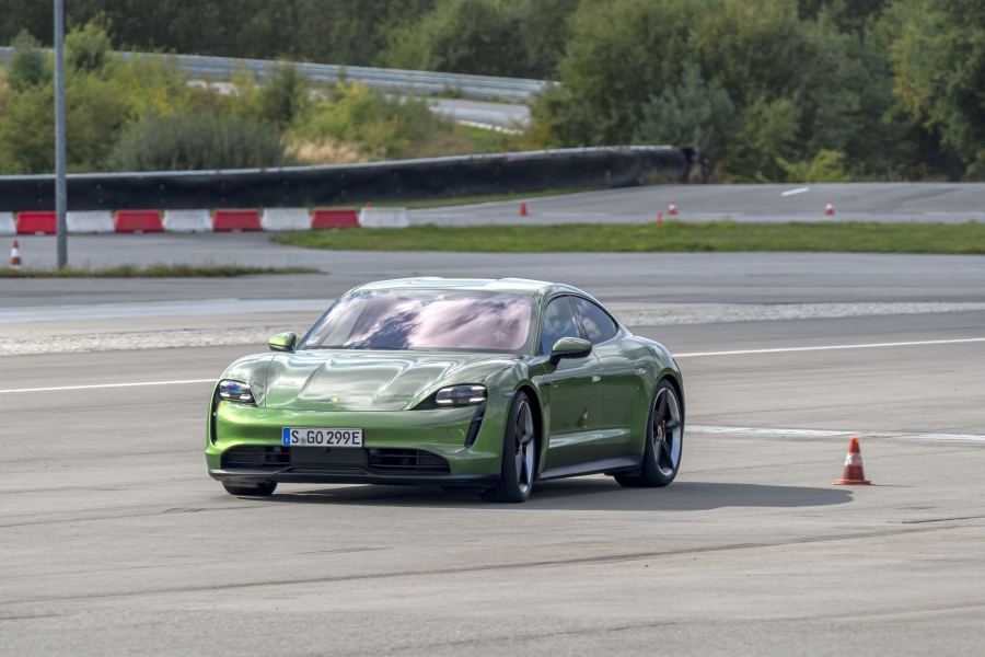 Zielone Porsche Taycan na torze mija pomarańczowy pachołek, a w tle widać drzewa i barierę.