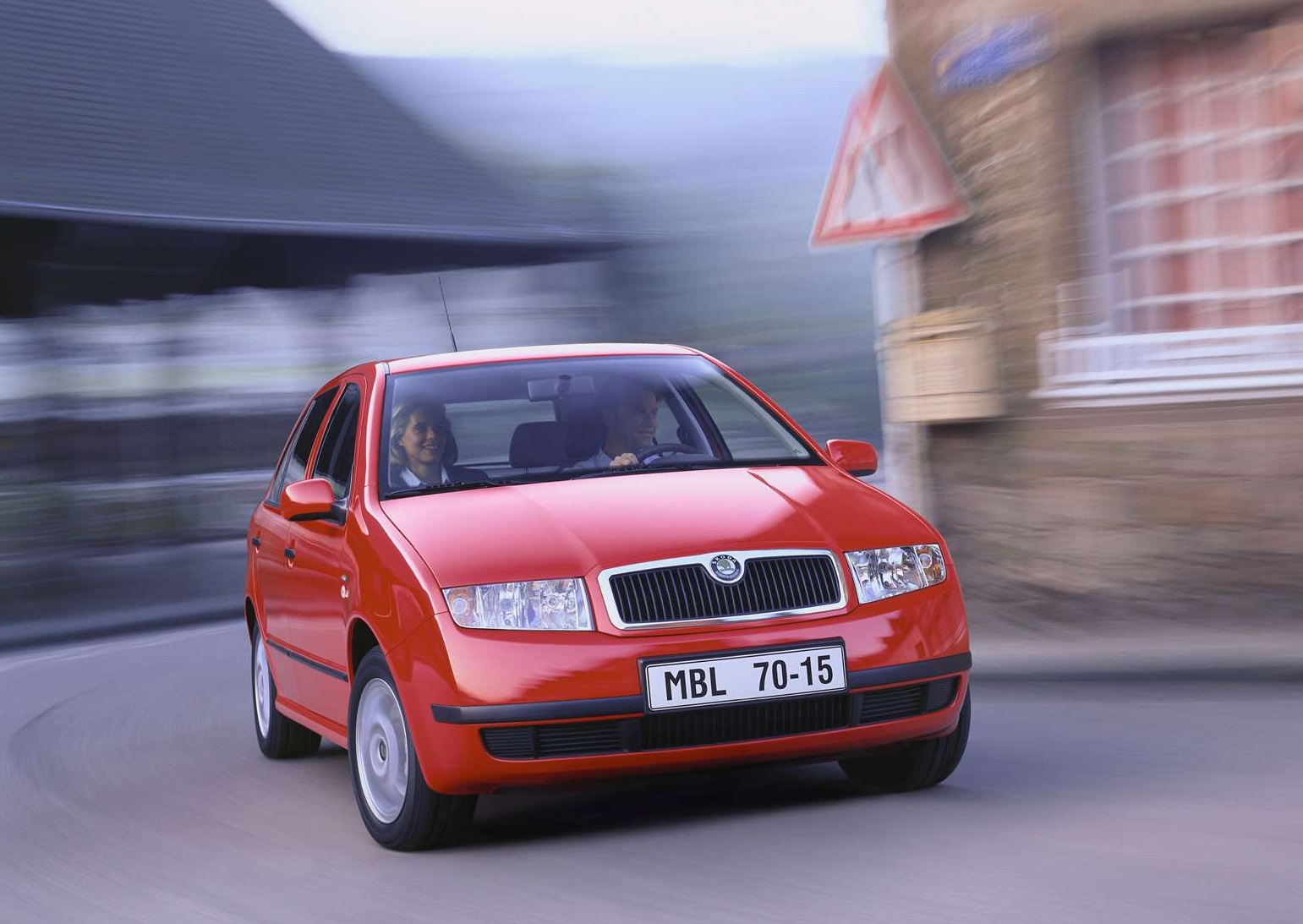 Czerwona Skoda Fabia z dwiema osobami jedzie po krętej ulicy w ruchu ulicznym.