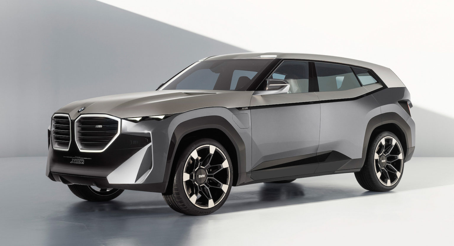 Srebrny prototyp SUV-a BMW XM stoi w jasnym, minimalistycznym studiu, w którym pojawiają się geometryczne cienie.