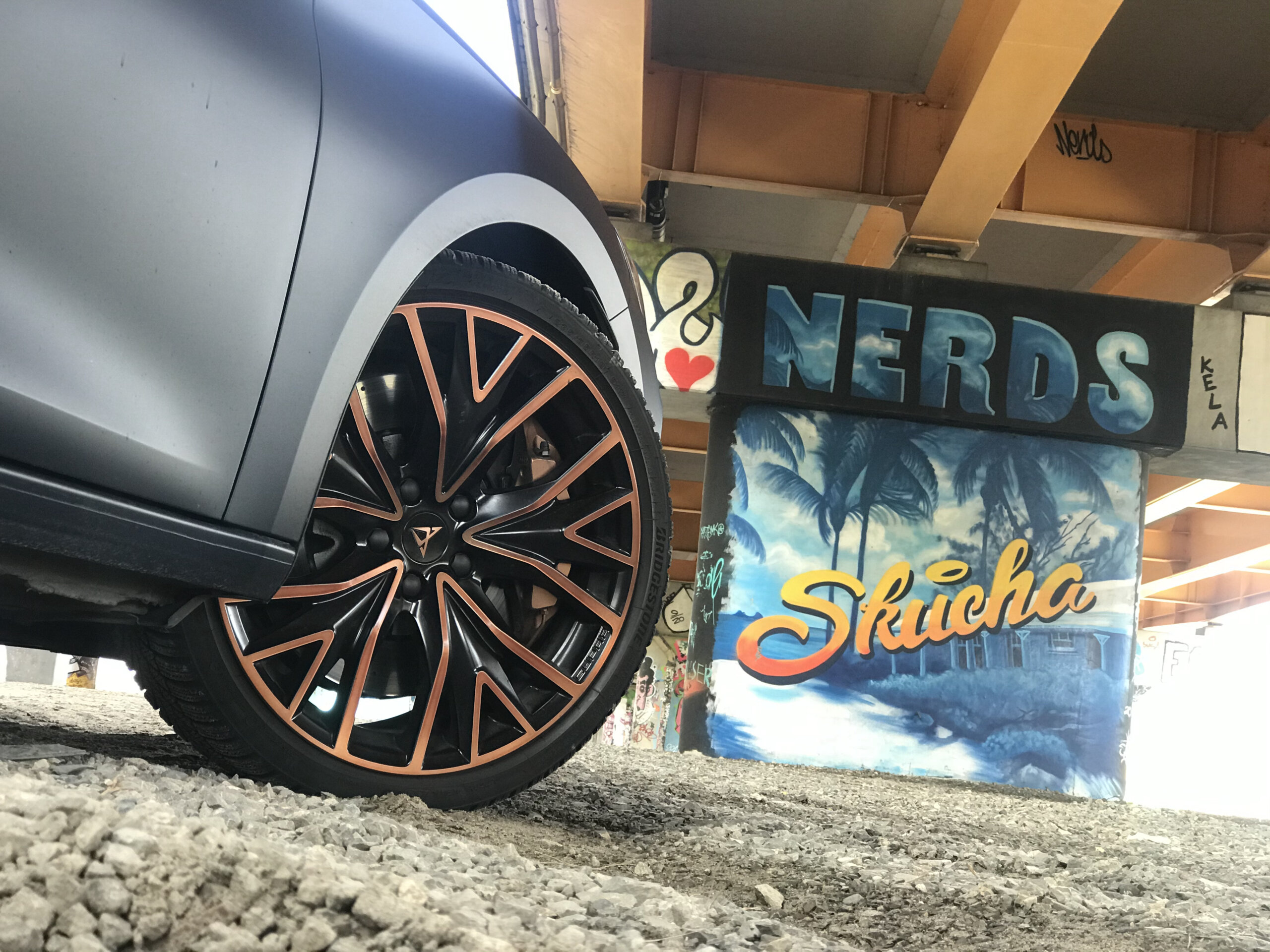 Cupra Leon ST z pomarańczowymi akcentami na żwirze, a w tle graffiti NERDS i Skucha.