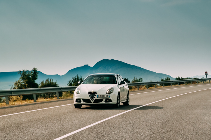 Białe Alfa Romeo Giulietta jedzie pustą, krętą drogą na tle gór i drzew.