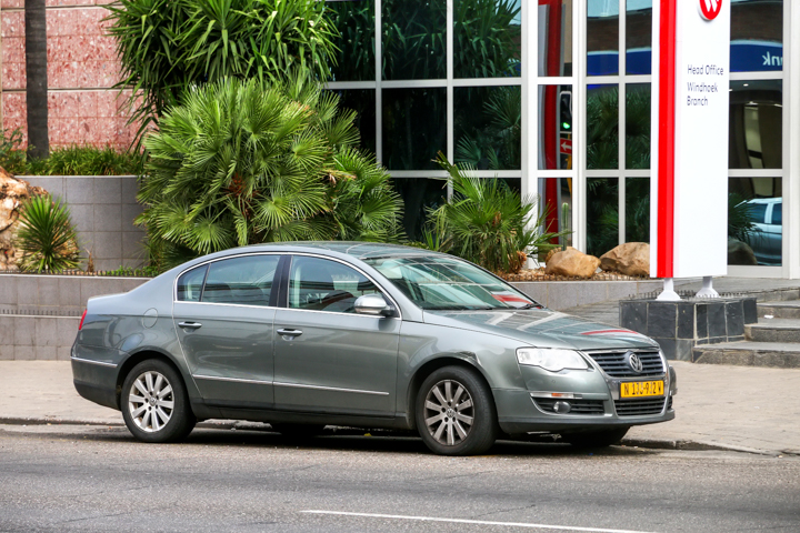 Szary Volkswagen Passat B6 z żółtą tablicą rejestracyjną stoi przy krawężniku obok budynku z palmami.
