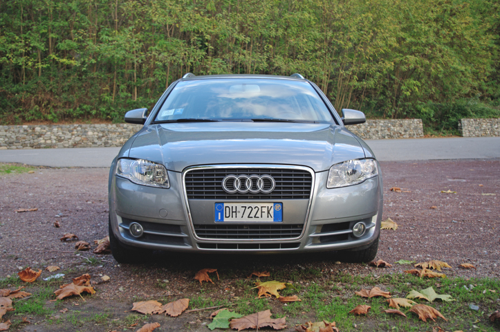 Szare Audi A4 B7 stoi na żwirze pokrytym liśćmi, przed drzewami i kamiennym murem.