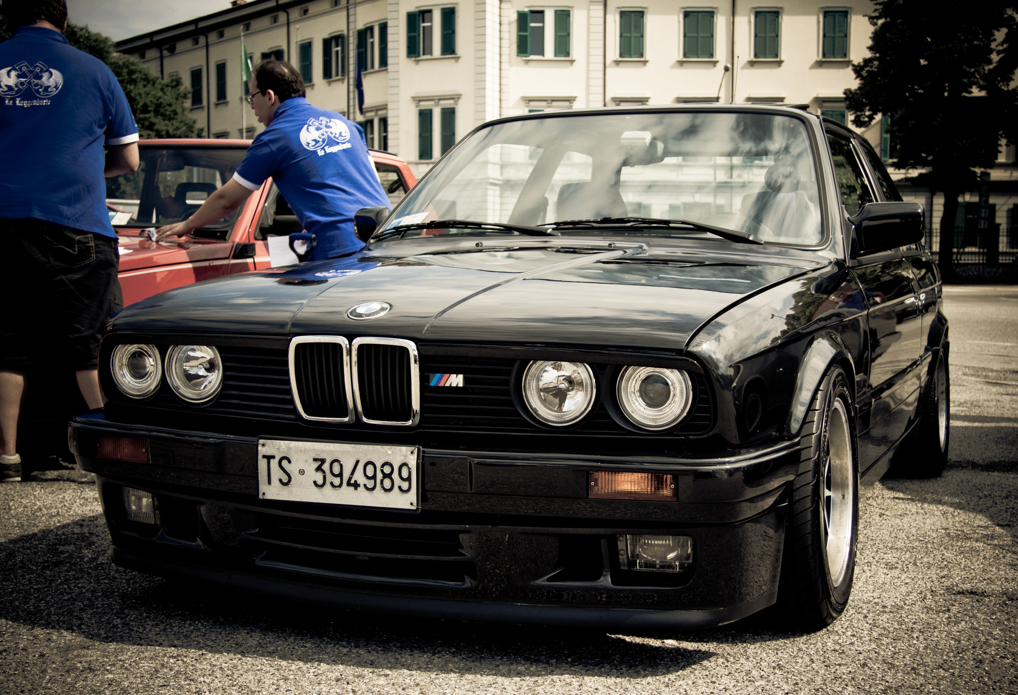 Czarne BMW E30 M3 z włoskimi tablicami rejestracyjnymi stoi na zewnątrz obok ludzi w niebieskich koszulach.