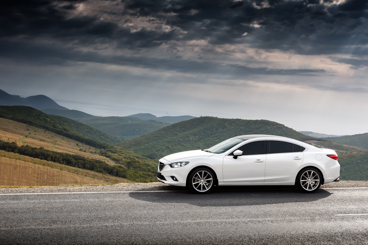 Biały sedan Mazda 6 stoi przy drodze, a w tle widać zielone wzgórza i góry pod chmurami.