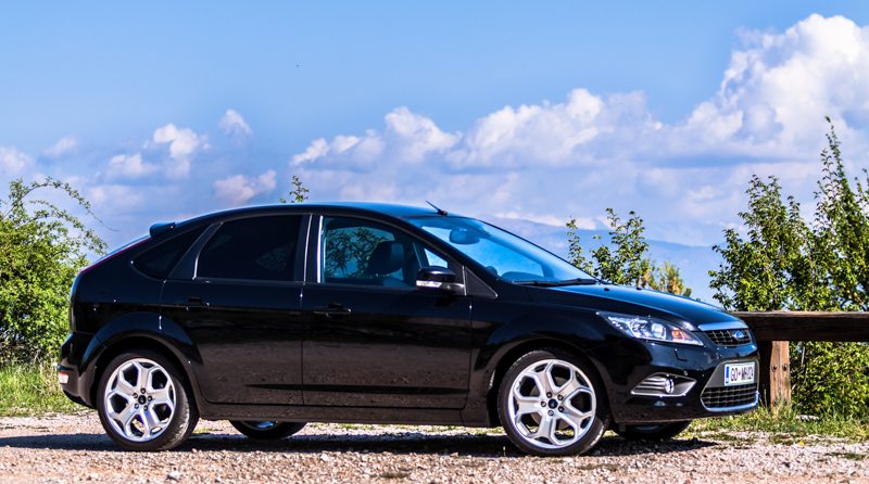 Czarny Ford Focus II w wersji hatchback stoi na żwirze, a w tle widać zielone krzewy i błękitne niebo.