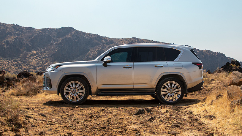 Srebrny Lexus LX stoi na suchym, kamienistym terenie, w otoczeniu wzgórz i pod błękitnym niebem.