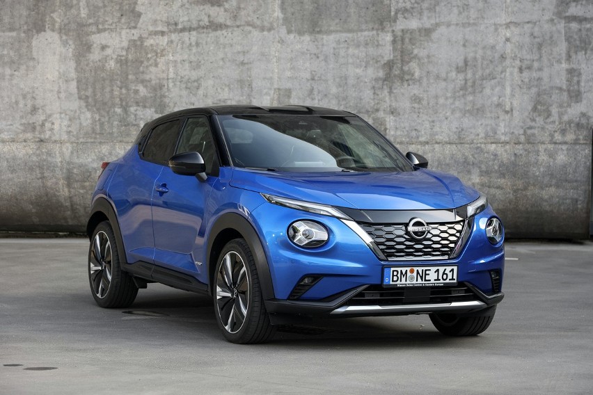 Niebieski Nissan Juke Hybrid stoi na betonie przed szarą, teksturowaną ścianą.