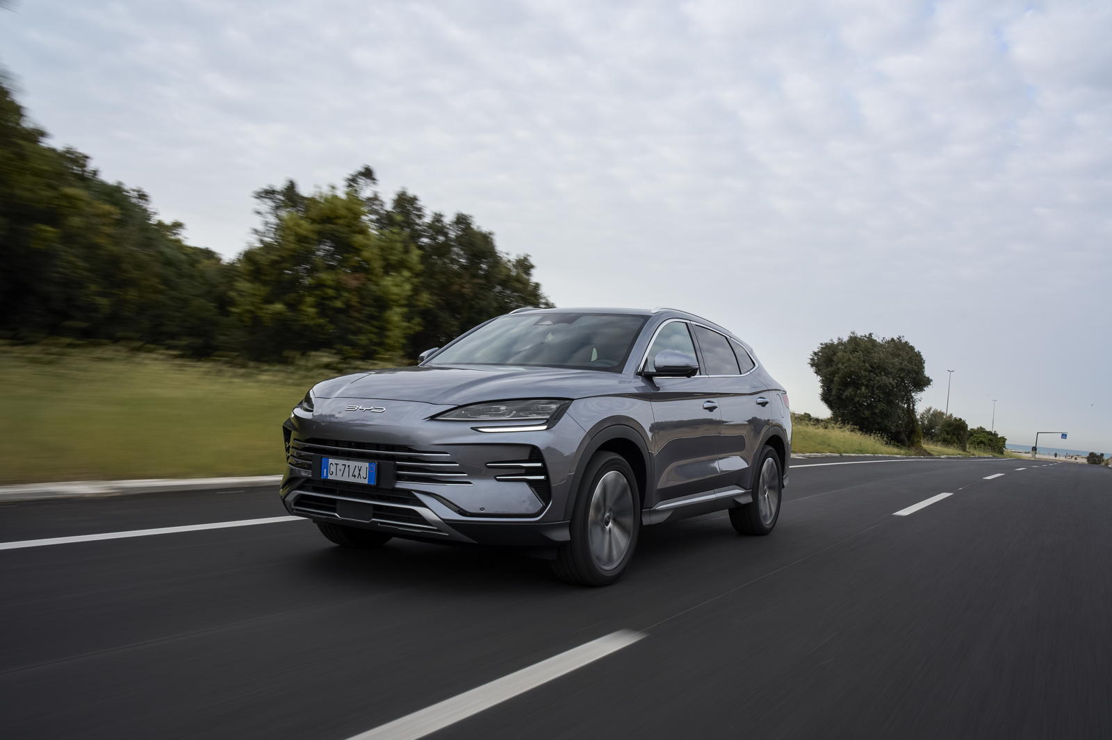 Srebrny SUV BYD Super DM jedzie po asfalcie obok drzew, trawy i pod chmurami.