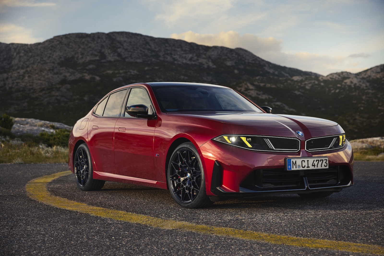 Czerwony sedan BMW zaparkowany na krętej górskiej drodze, w tle wzgórza i niebo.