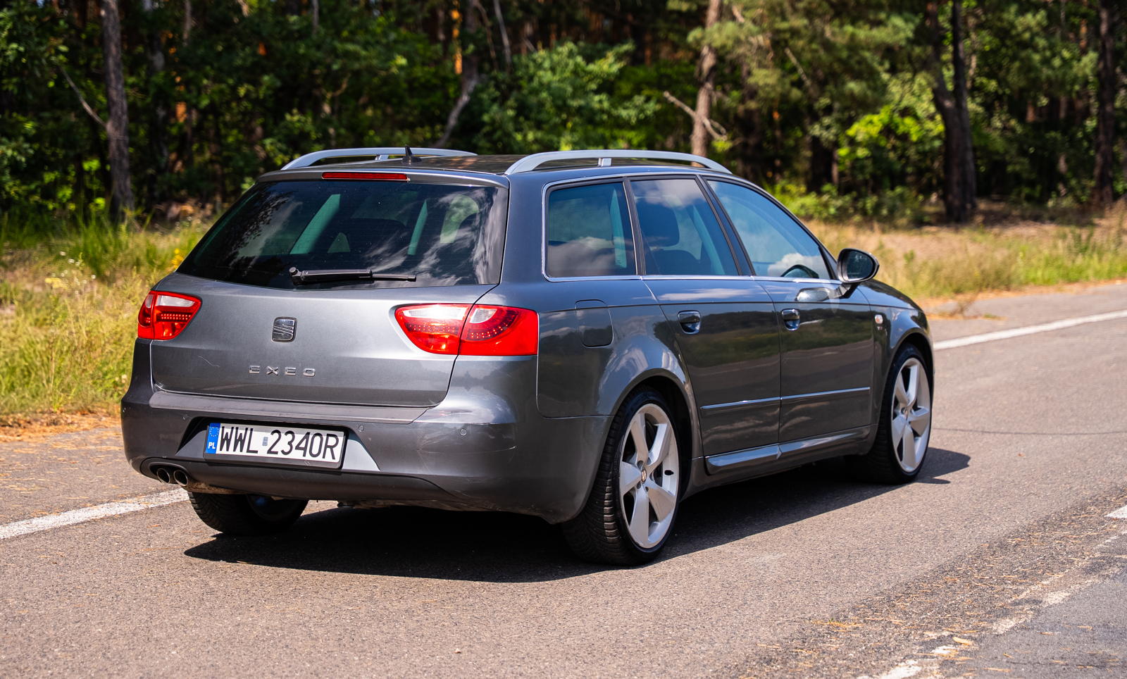 Szary, używany Seat Exeo kombi stoi przy drodze obok lasu w słoneczny dzień.