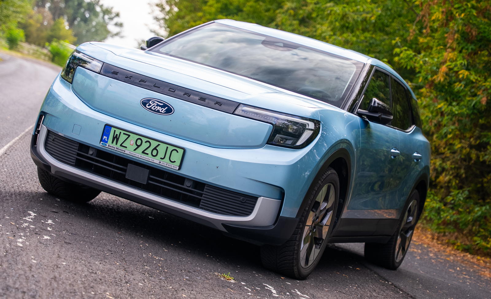 Niebieski Ford Explorer EV z numerem rejestracyjnym WZ 926JC stoi przy drodze obok drzew.
