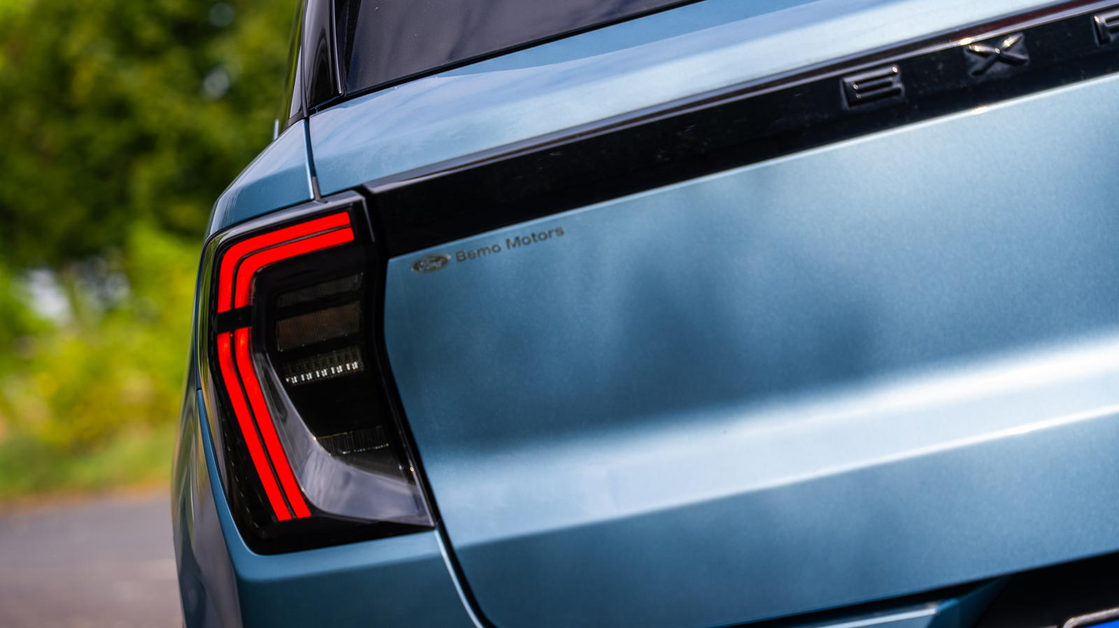 Lewy tylny reflektor i fragment bagażnika niebieskiego samochodu z logo Bano Motors.