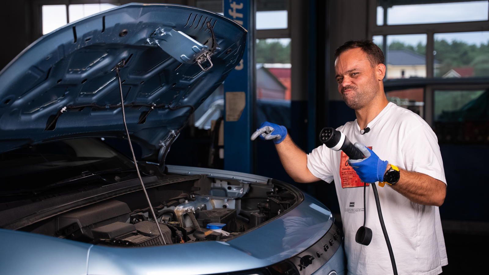 Mężczyzna w rękawiczkach trzyma wtyczkę ładowarki do samochodu elektrycznego obok używanego Forda Explorera z otwartą maską.
