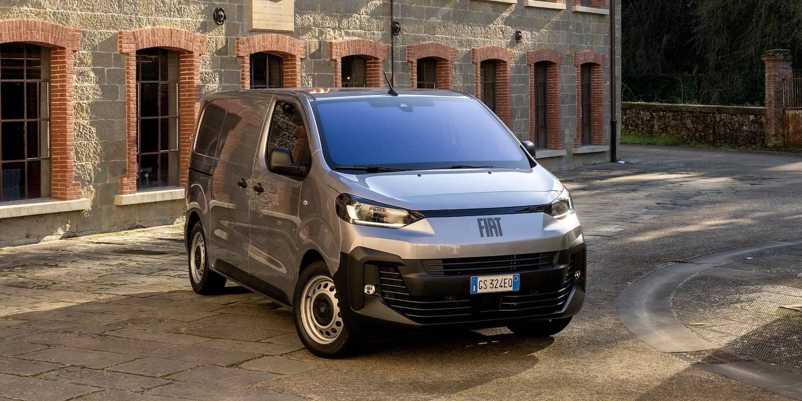 Srebrny van marki Fiat stoi na kamiennym podjeździe przed budynkiem z cegły i kamienia.