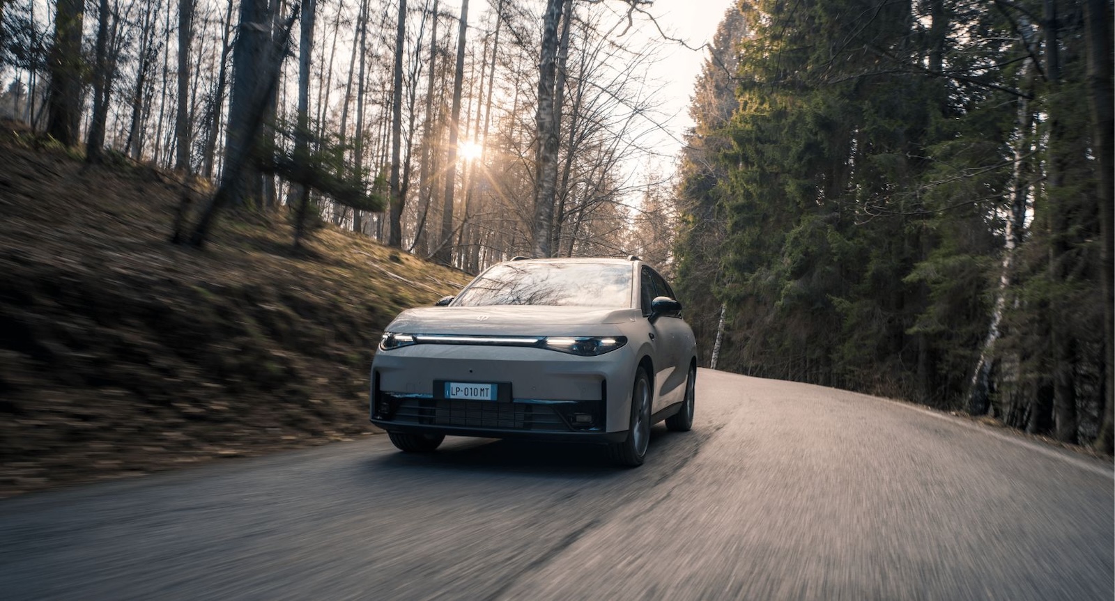 Srebrny Leapmotor C10 jedzie leśną drogą, a promienie słońca przebijają się przez drzewa.