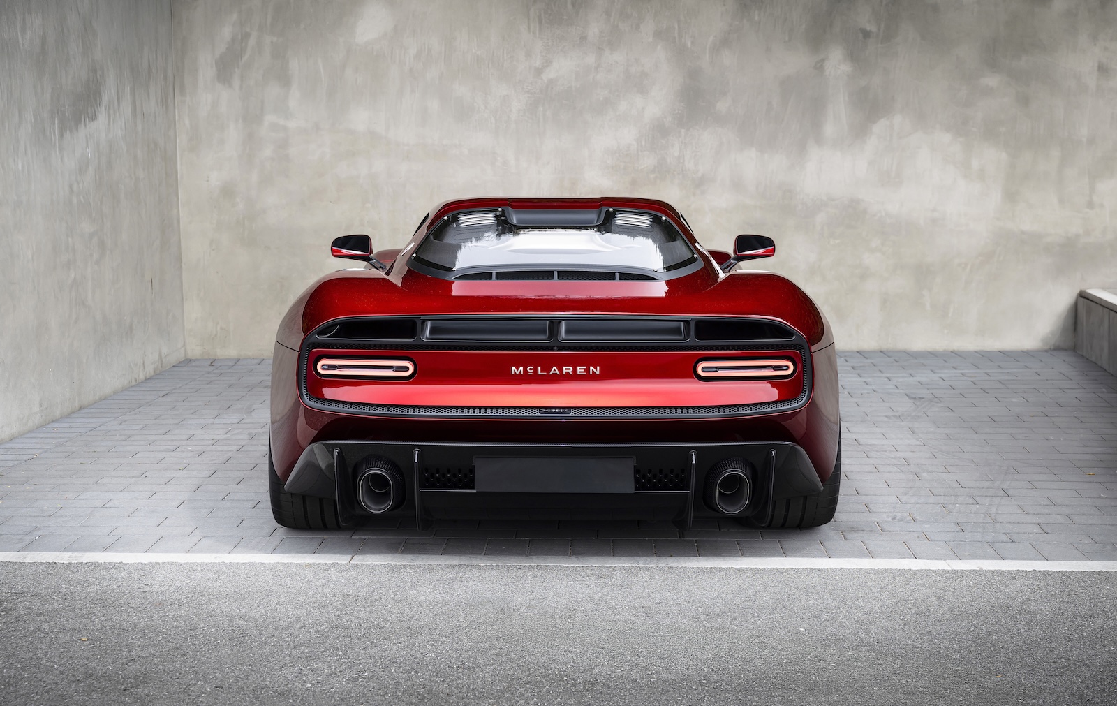 Czerwony McLaren widziany od tyłu na szarej kostce, na tle betonowej ściany.