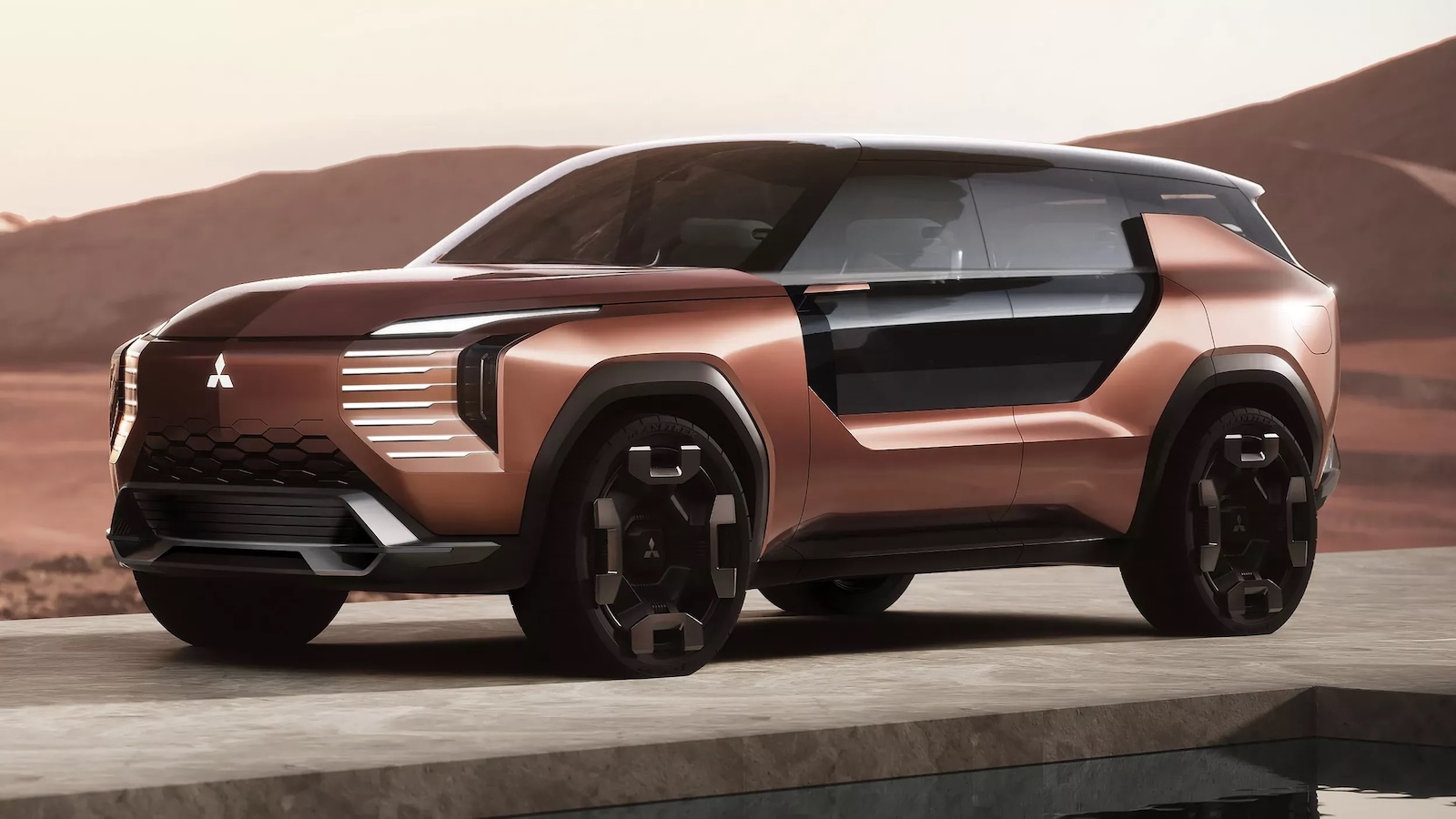 Brązowy prototyp SUV-a marki Mitsubishi stoi nad wodą, a w tle widać góry i zamglone niebo.
