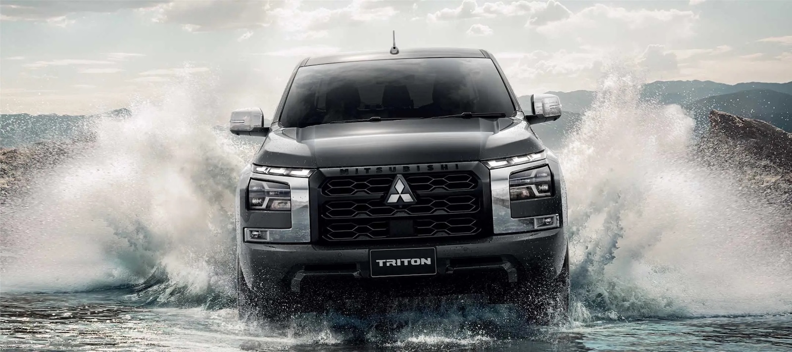Mitsubishi Triton jedzie przez wodę, rozpryskując ją, a w tle widać góry i chmury.