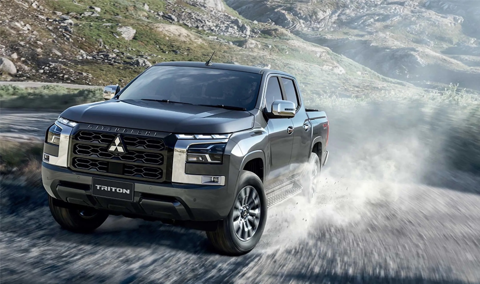Mitsubishi Triton jedzie po zakurzonej, kamienistej drodze w górzystym terenie.