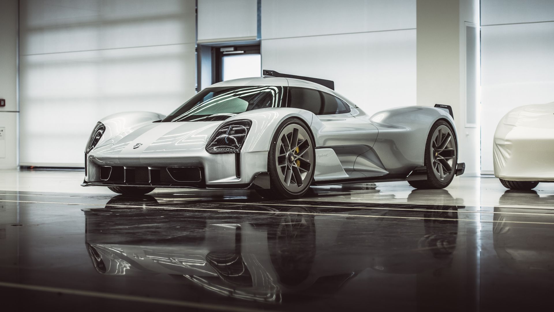 Srebrne Porsche na lśniącej podłodze obok zakrytego samochodu w jasnym salonie.