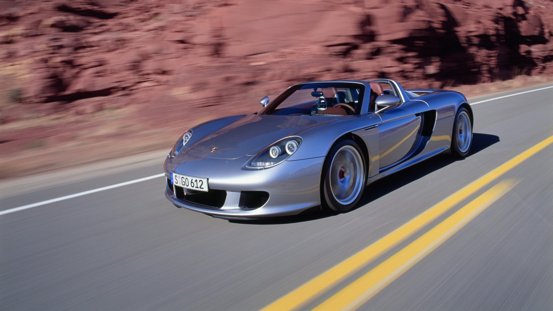 Srebrne Porsche Carrera GT jedzie drogą na tle czerwonych skał.